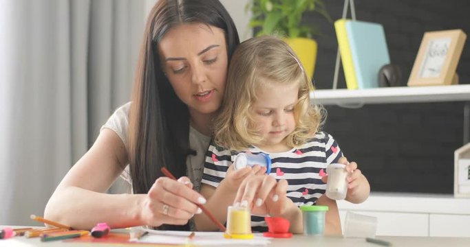 Pretty little girl with blond curly hair opening box with plasticine and her mother with black long hair drawing using pencil at home. Indoor.