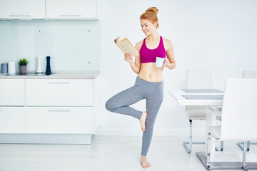 Fototapeta na wymiar Portrait of fit red haired woman doing yoga exercises at home and reading book, smiling