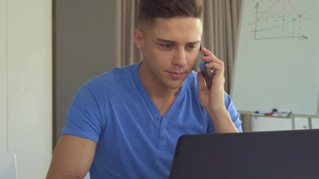 Attractive brunette guy answering the call at the office. Close up of handsome male manager sitting at the desk with laptop. Cute young man using smartphone at his work