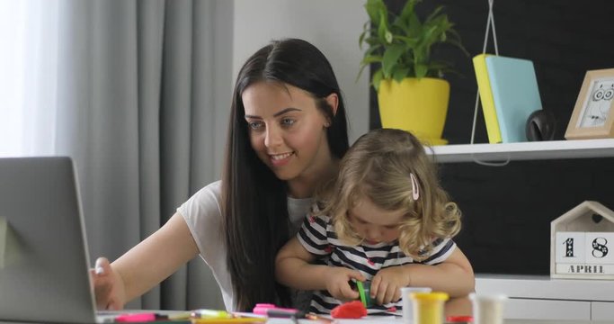Attractive caucasian young woman with black long hair working on laptop while her dauther opening box with plasticine at home. Indoor.