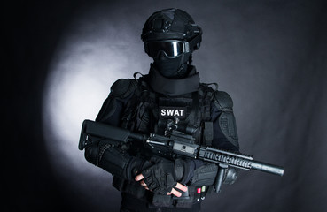 Member of the SWAT squad with an assault rifle in a black uniform on dark background. Special weapons and tactics. Special Forces. - obrazy, fototapety, plakaty