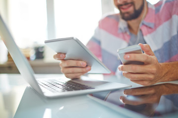 Close-up of young freelancer using modern devices to receive information, he sitting at table with laptop and holding phone and tablet in hands