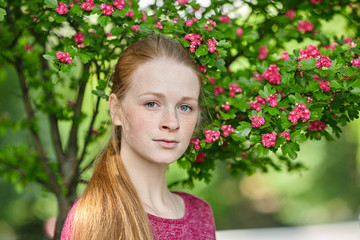 Fototapeta premium Closeup portrait of young natural beautiful redhead woman in fuchsia blouse posing against blossoming tree with blurred green foliage park background