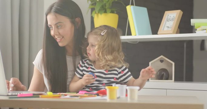 Caucasian pretty girl with blond curly hair opening box with plasticine while her mother using laptop at home. Indoor.