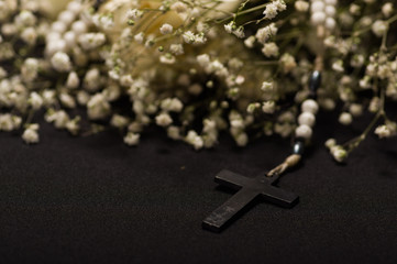 Close up of a rosary beads with a black cross in blurred white small flowers, black background