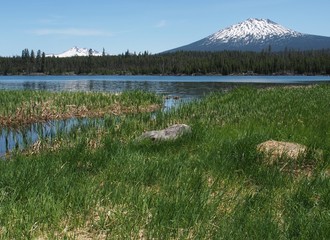 The calm blue waters of Lava Lake with a channel running through green reeds and Mt. Bachelor and Broken Top in the background in Oregon's Cascade Mountains on a sunny summer day.  - Powered by Adobe