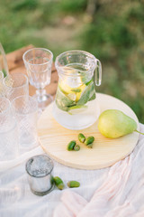 weekends, party, celebration, wedding, serveware concept - transparen pitcher with iced mojito nearby with wine-glasses and yellow-green pear on light wooden board