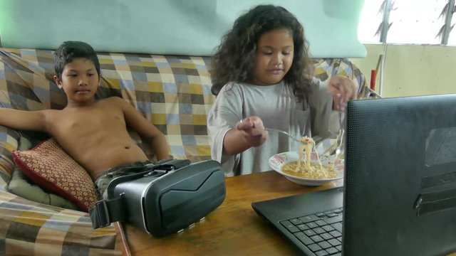 Medium shot multicultural Asian siblings watch a video while the sister eats a bowl of noodles;  a Virtual reality VR headset rests on the table nearby / One of a series