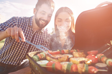 Friends making barbecue and having lunch in the nature. Couple having fun while eating and drinking at a pic-nic - Happy people at bbq party - obrazy, fototapety, plakaty