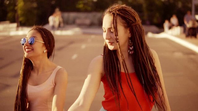 Two happy women with dreads walking on the empty road and talking in summer. Two hipster girls laughing and dancing during a bright sunny day. Slowmotion shot.