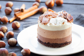 Mousse cake three chocolate, whole hazelnut and cinnamon sticks on a white plate. Dark wooden background