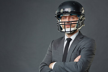 Confident businessman wearing suit and American football helmet standing cross-armed against grey...