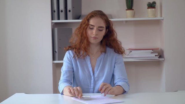 Portrait successful business woman with curly redheaded long hair sitting at the white desk in office sign document. Attractive female wearing casual elegant blue shirt undersign contract looking at