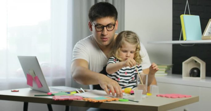 Caucasian little girl with blond curly hair using paints for drawing on a sheet of paper siting with her father in home.