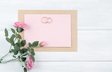 Pink roses and bridal rings with paper greeting card for wedding on background of shabby wooden planks.