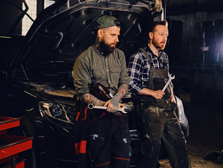 Two bearded tattooed mechanics near the car in a workshop.