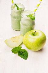 Freshly blended green apple fruit smoothie in glass jars with straw, mint leafs, apples. White wooden board background, copy space.
