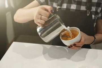 Barista pouring cream from a jug into a Cup of coffee, holding them in his hands, standing in the sun, closeup photo