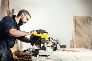 Young man builder carpenter sawing board with circular saw in workshop