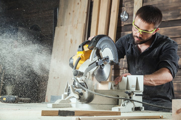 Close-up as a young male construction worker carpenter saws a circular saw blade, sawdust fly in the workshop - obrazy, fototapety, plakaty
