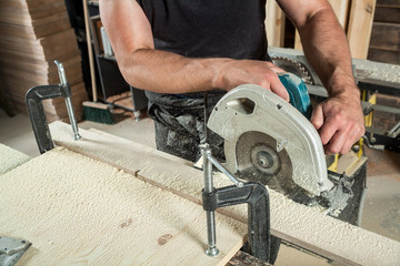 A young man saws a wide wooden board with a circular saw in the workshop