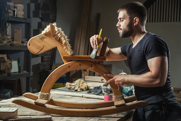 A young man paints a wooden toy with a paint brush with a protective varnish, made by himself from...