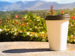 White cup of coffee put on rock table on the rose garden with blurred mountain and gold light sky and clouds background in the morning of Chart Farm, Cape Town, South Africa