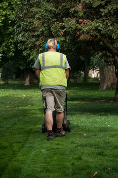 jardinier de dos avec un casque anti-bruit tondant la pelouse dans un jardin public