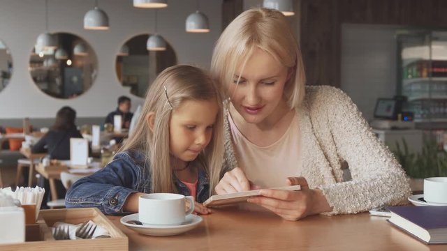 Pretty caucasian mother and daughter looking at the menu. Attractive blond woman asking her child what she wants to drink at the cafe. Cute little girl pointing her forefinger on board
