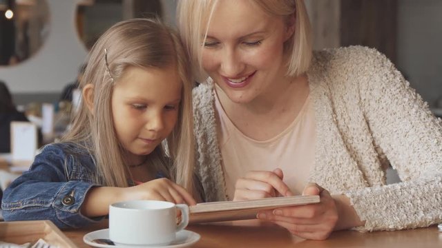 Attractive blond mother and her daughter choosing drinks at the cafe. Close up of pretty young woman holding menu in her hand. Cute little girl showing her mother what she wants to drink