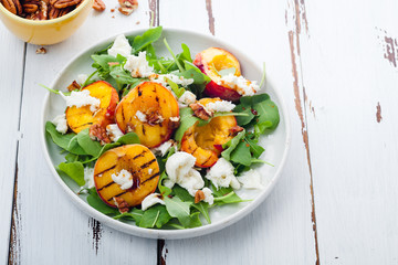 Fresh salad with grilled peach halves, arugula and burrata on a plate on white distressed wooden background. Top view, horizontal composition with copy space. Summer food concept
