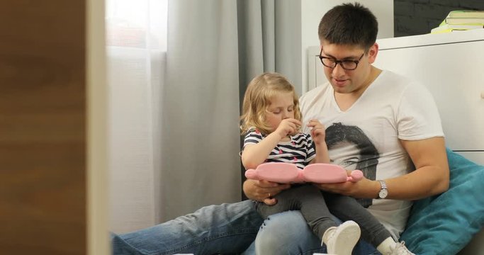 Young caucasian man in glasses siting on the chair in home with his charmind daughter, using tablet for looking images . Indoor.