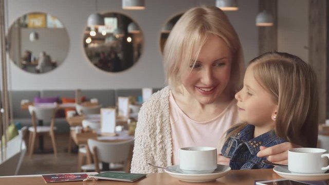 Attractive little girl sharing some secrets with her mother at the cafe. Prety young woman wondering at what her daughter whispering. Cute blond mom kissing her daughter at the table
