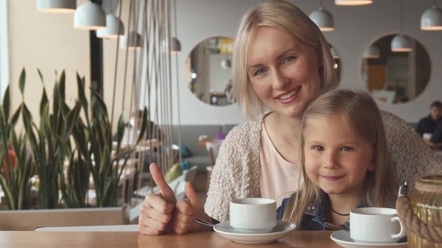 Pretty blond mother and her daughter showing their thumbs up at the cafe. Attractive young woman and her female child approving restaurant. Little caucasian girl and her mom smiling for the camera