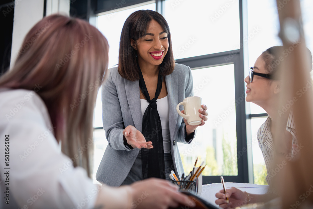 Wall mural smiling young businesswomen talking and drinking coffee in office