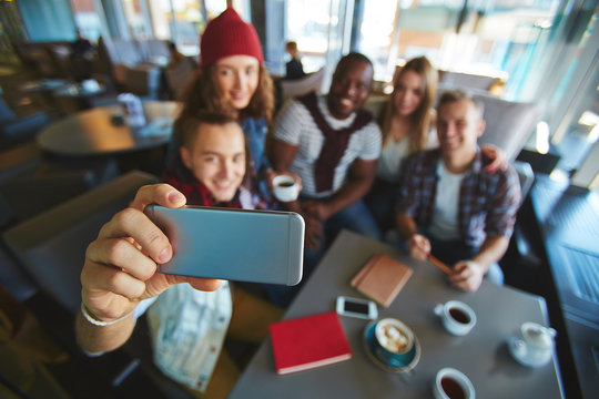 Group of friends taking selfie on smartphone while gathered together in lovely coffeehouse with panoramic windows, focus on foreground