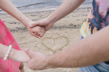 A young couple holds hands by the river. In the sand, a heart is drawn