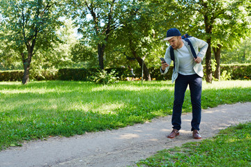 Full length portrait of handsome young man walking in sunny public park with backpack and browsing...