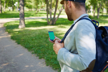 Unrecognizable man with backpack using smartphone while walking in sunny morning park