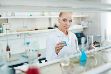 Fototapeta premium Portrait of beautiful female chemist working in modern laboratory checking test tubes and looking at camera behind shelves with glassware