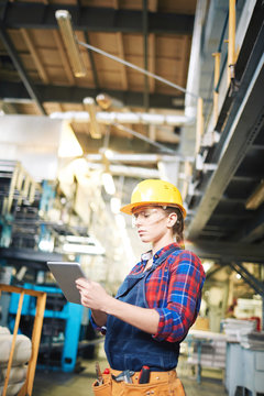 Profile view of confident female technician in protective eyewear and hardhat using digital tablet in modern factory, waist-up portrait