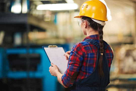 Back view of female warehouse worker wearing protective goggles and helmet taking inventory with clipboard, blurred background