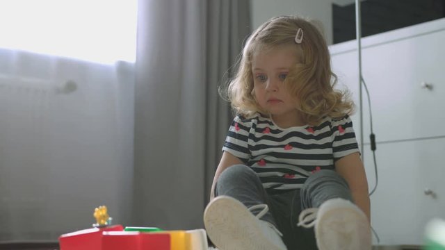 Beautiful little girl with blond curly hair siting on the floor and showing on her toys on living background. Indoor.