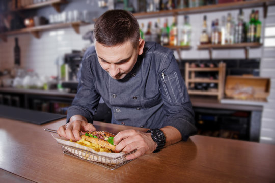 Cheerful chef in uniform, laying out a delicious hamburger with fried potatoes in the basket.