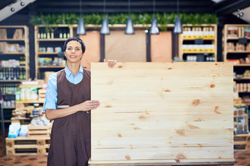 Pretty middle-aged shop assistant in apron holding empty wooden board while standing in lovely store with organic products, portrait shot