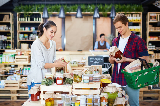 Young Couple Reading Labels On Jars While Doing Shopping In Local Store With Farm Products