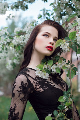 Fototapeta premium Fashion art Woman in summer near blossoming Apple tree, bright lipstick. Spring summer mood, mysterious romantic image of girl in a black dress tree flowers. Woman dreams