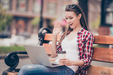 Hello there! Young girl is waving to the screen to her boyfriend in camera while having video call in the spring park outdoors, sitting on the bench with hot tea