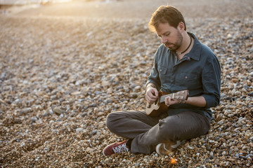 Handsome man playing ukulele in a stone beach during sunset