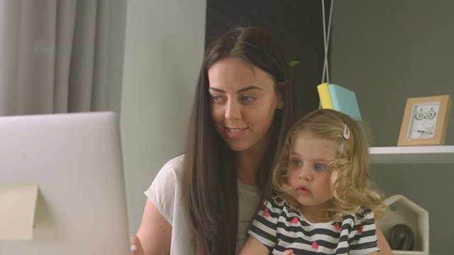 Attractive caucasian woman with long black hair working on the laptop and her charmind little dauther with blond curly hair sculpturing with plasticine on living background. Indoor.